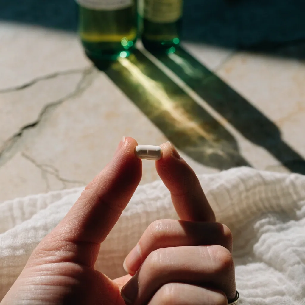 A matte white capsule held between two fingers above a folded muslin cloth under raking noon light